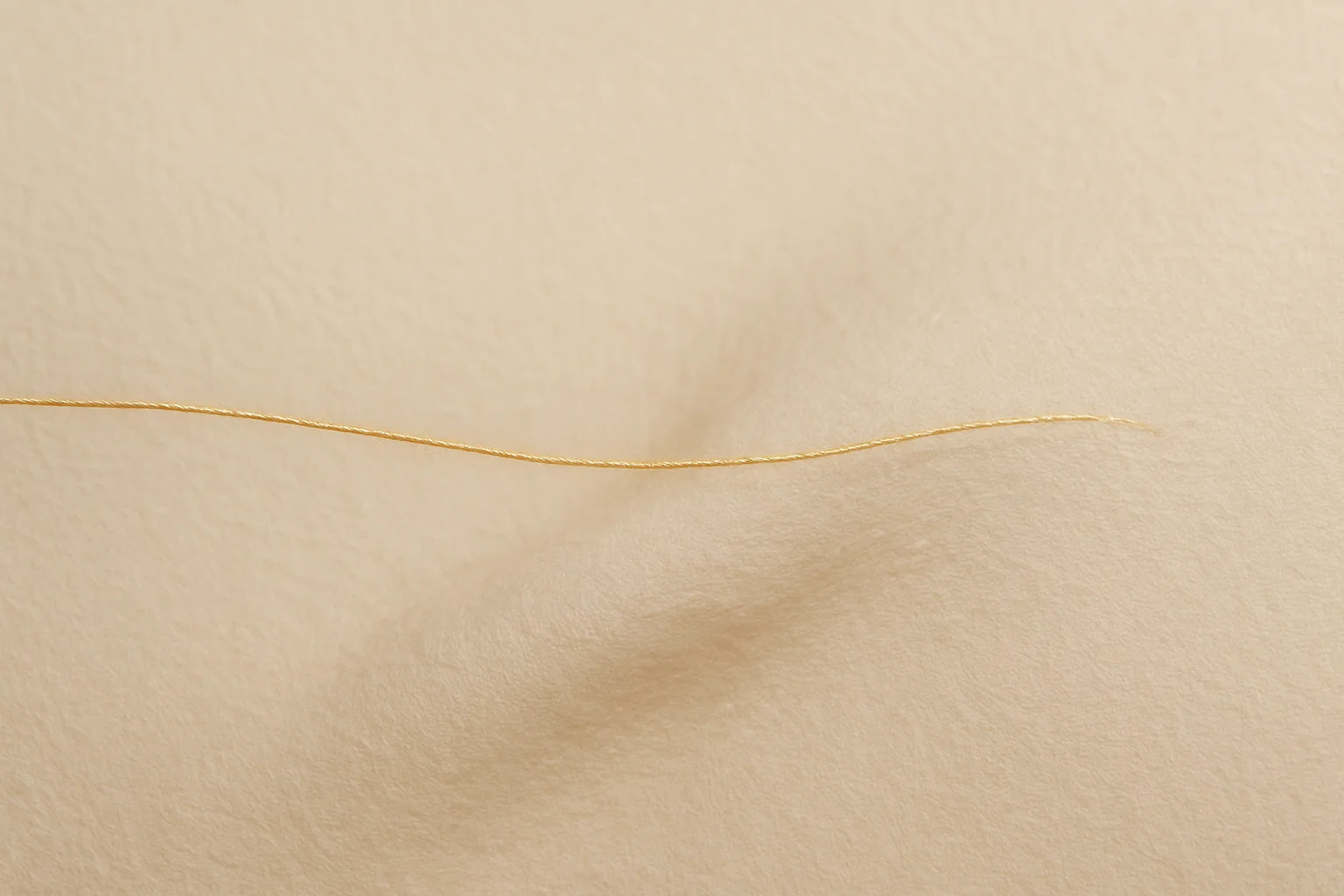 Ein einzelner, zarter Goldfaden (#ccb974) spannt sich über eine weiche, beige Fläche (#fcf8ef), die wie Haut wirkt. Die minimalistische Komposition strahlt leise Spannung und fragile Stärke aus.